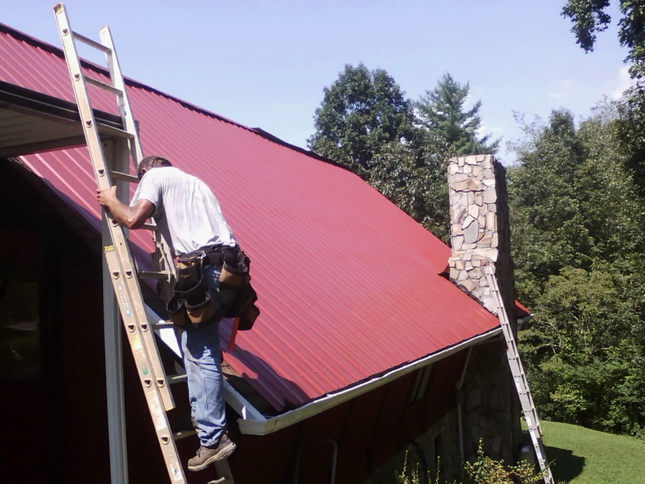 Roofer on a ladder beside a red metal roof and stone chimney — Soffit & Fascia Repair by Roofing Brothers in Perryopolis